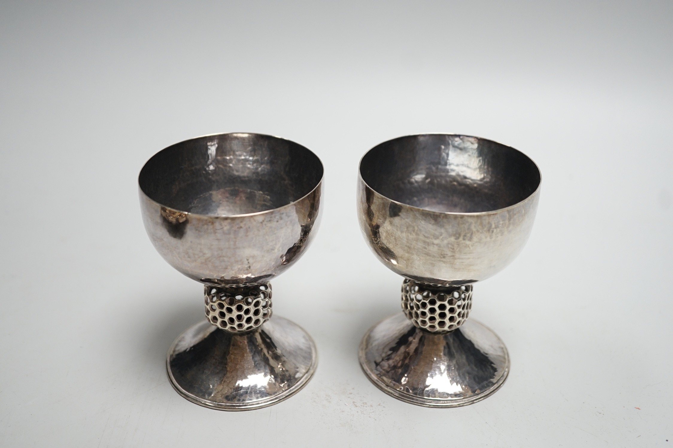 A stylish pair of Elizabeth II Arts & Crafts style planished silver pedestal cups, with reticulated stems, by Oliver Bailey, London, 1973, height 93mm, 7.5oz.